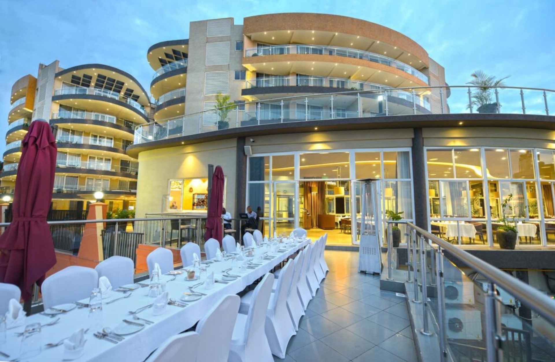 Skyz Hotel Kampala's curved building at dusk, with a long white-clothed banquet table set on Aruba Terrace