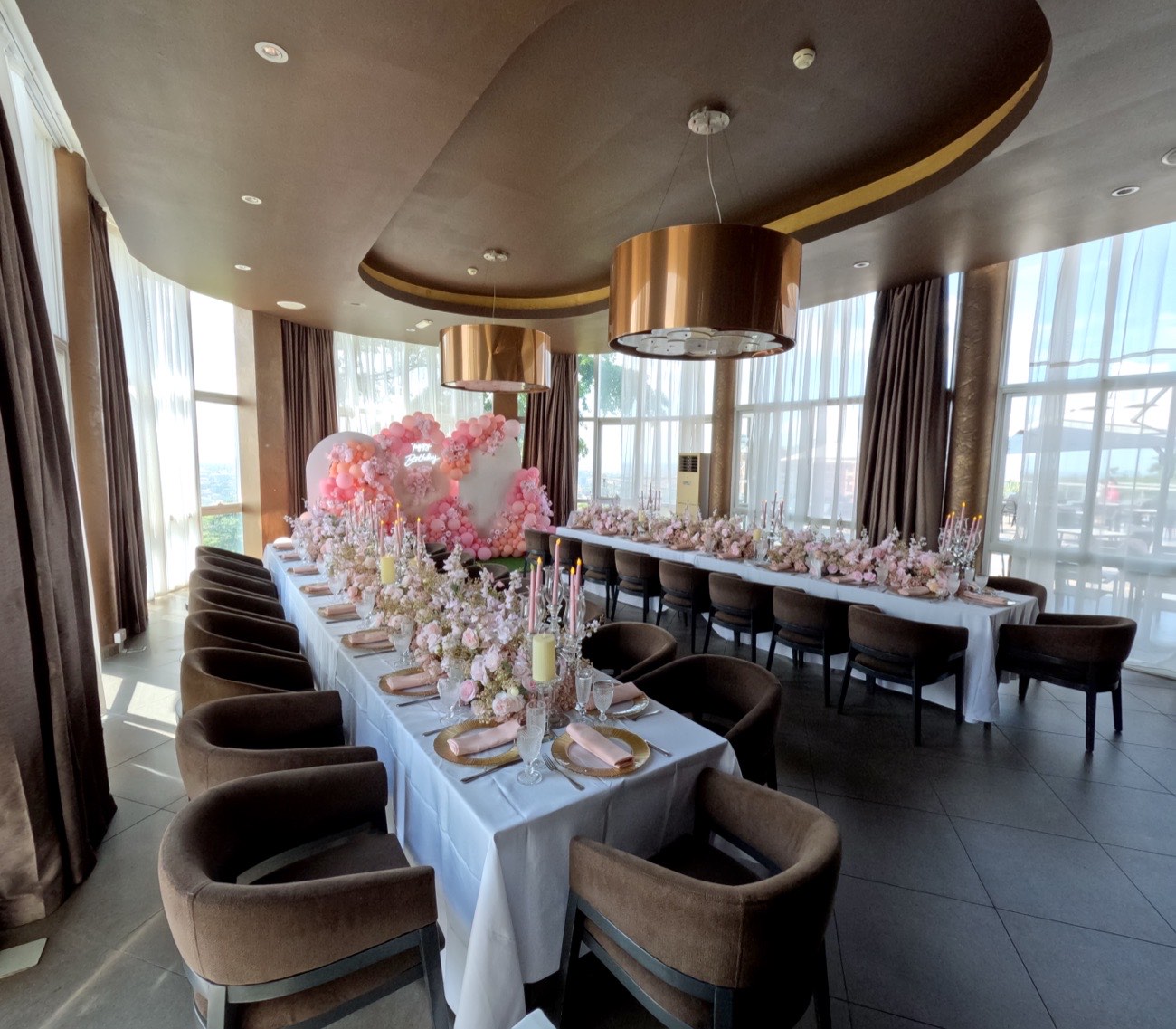 Long guest table dressed with blush floral arrangements and pink taper candles inside the VIP Room at Skyz Hotel Kampala, tall windows framing the city skyline