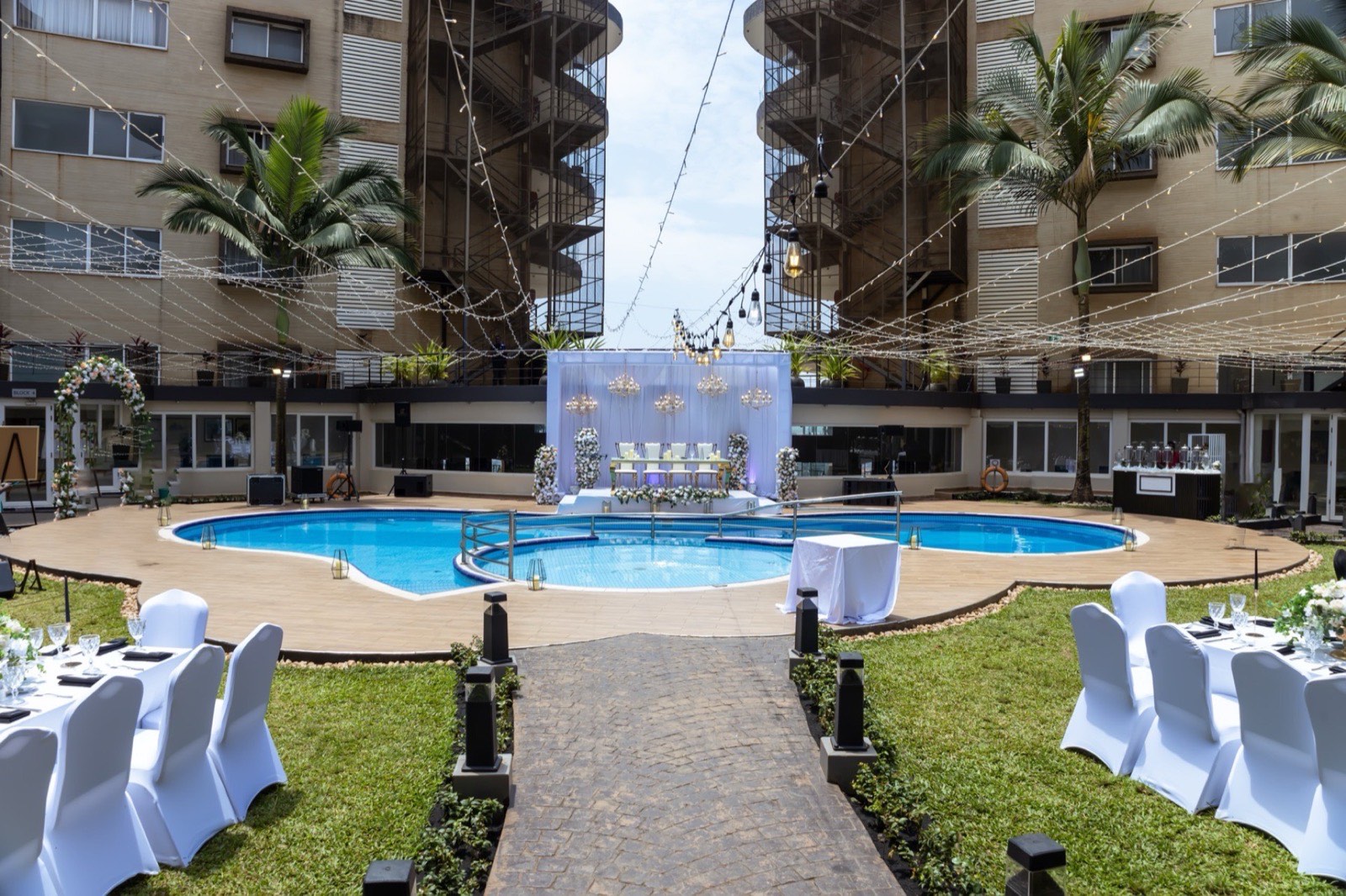 Aerial view of a wedding reception around Skyz Hotel Kampala's twin pool courtyard by day, framed by palm trees and string lights